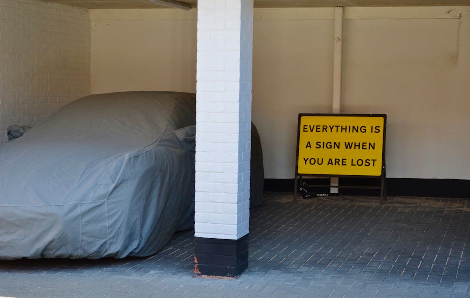 The image shows a partially covered car parked in an indoor or enclosed garage area, with a light grey or silver-grey fabric cover draped over it, exposing only the shape and contours of the vehicle's roof and part of the hood. The garage has white-painted brick walls, with a central white brick column supporting the structure, and a dark, textured floor made of small, rectangular tiles arranged in a herringbone pattern. To the right of the covered car, there is a yellow and black sign on a portable stand with the message 