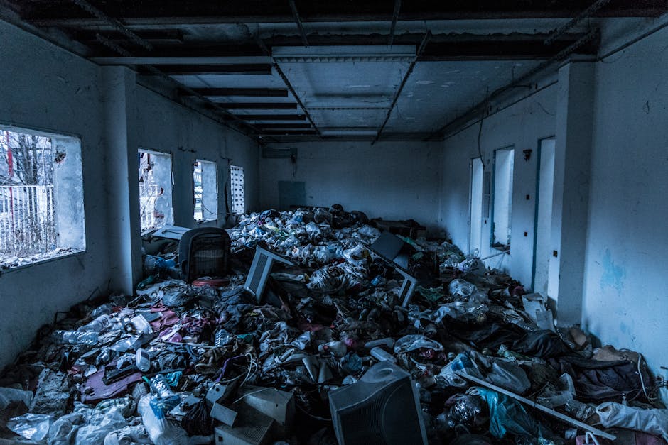 The image shows a large indoor room with white walls and a ceiling exposing structural beams and electrical wiring. The floor is completely covered with a substantial pile of discarded waste materials, including tangled plastic bags, cardboard boxes, electronic devices such as monitors and speakers, and various unidentifiable rubbish. Several of the waste items are partially crushed or torn, with some objects leaning or stacked haphazardly amidst the mess. Natural light enters through multiple rectangular windows along the left wall, revealing a view of trees and a metal fence outside. The room appears to be in a state of neglect or abandonment, with no furniture or cleaning equipment visible. The scene closely relates to private waste disposal or on-site clearance activities, potentially indicating a situation where independent rubbish removal services, like those offered by Rubbish Removal Holland Park, are needed to clear accumulated debris from an interior space.