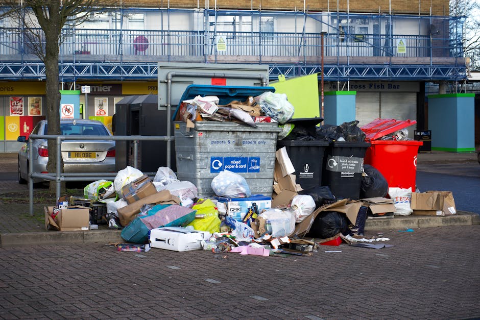 The image shows a large indoor room with white walls and a ceiling exposing structural beams and electrical wiring. The floor is completely covered with a substantial pile of discarded waste materials, including tangled plastic bags, cardboard boxes, electronic devices such as monitors and speakers, and various unidentifiable rubbish. Several of the waste items are partially crushed or torn, with some objects leaning or stacked haphazardly amidst the mess. Natural light enters through multiple rectangular windows along the left wall, revealing a view of trees and a metal fence outside. The room appears to be in a state of neglect or abandonment, with no furniture or cleaning equipment visible. The scene closely relates to private waste disposal or on-site clearance activities, potentially indicating a situation where independent rubbish removal services, like those offered by Rubbish Removal Holland Park, are needed to clear accumulated debris from an interior space.