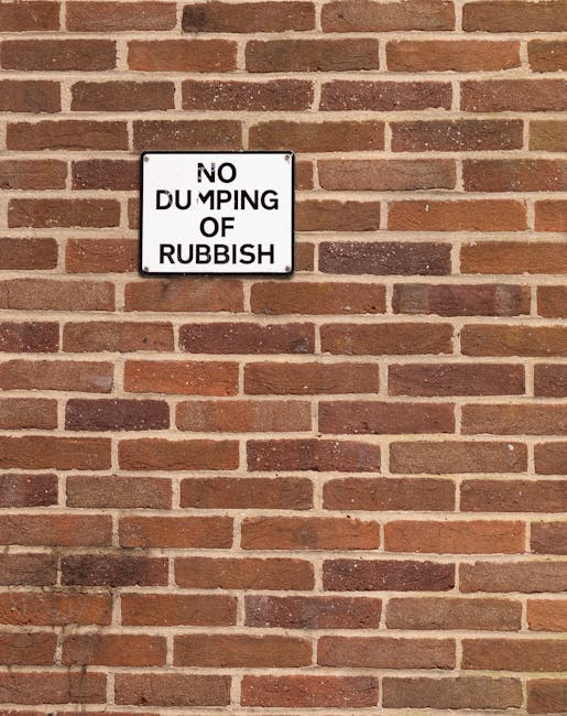 A rectangular metal sign mounted on a red-brown brick wall, displaying the message 'NO DUMPING OF RUBBISH' in black capital letters on a white background. The sign is positioned centrally within the image, with the brick wall extending evenly around it. The wall features a traditional running bond pattern with bricks of varying shades of red, brown, and a few darker tones, separated by light grey mortar joints. The surface of the wall appears clean and well-maintained, with a slightly matte finish. The lighting is natural and diffuse, casting soft shadows that highlight the brick texture and the flat surface of the sign. The scene suggests a location where waste disposal is monitored, possibly indicating an area under private management or a designated rubbish collection point. The presence of the sign reflects efforts to regulate rubbish disposal and prevent illegal dumping, aligning with themes of private waste handling and alternative rubbish removal services like those offered by Rubbish Removal Holland Park.