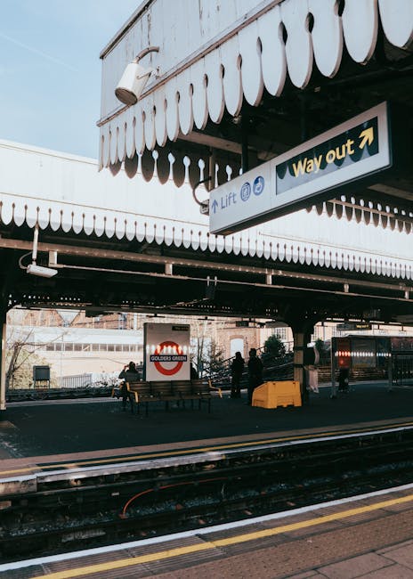 A view of Holland Park Station platform featuring a flat, textured black canopy with a scalloped white trim along its edge, supporting a rectangular overhead sign indicating directions to the lift and way out. The sign has a black background with white and yellow lettering and directional arrows. In the background, there are a few seated and standing passengers, some standing near a yellow safety barrier, which appears to be a low, sturdy, plastic construction for crowd control or safety purposes. Behind the platform, railway tracks extend into the distance, with metal rails and dark sleepers visible. Further back, a large sign displaying the London Underground roundel with the words 'GOLDERS GREEN' is mounted on a white wall or glass panel, part of the station infrastructure. The overall environment is well-lit by natural daylight, casting shadows on the platform, and the scene is typical of a busy urban railway station, with elements supporting everyday travel and independent transportation options, including elements of on-site clearance for maintenance or rubbish disposal at the station.