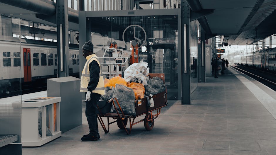 A worker wearing a dark jacket, light safety vest, gloves, and a beanie hat stands on a modern train station platform beside a red hand truck loaded with black, grey, and orange garbage bags filled with waste. The bags are stacked and spilling slightly, indicating they are used for rubbish collection, with some appearing crumpled or tied. The platform has a smooth, grey tiled surface, and behind the worker, there is a glass-enclosed lift or elevator with metallic framing, reflecting the station’s architecture. To the left, a white waste bin with an open top is visible, possibly designated for general rubbish. In the background, an overhead train with closed doors is parked at the platform, and a few passengers are seen walking further along the station. The environment is well-lit with natural daylight, highlighting the utilitarian, urban setting. This scene underscores private waste handling and manual rubbish removal from public transport hubs, illustrating methods of on-site clearance relevant to services offered by Rubbish Removal Holland Park.