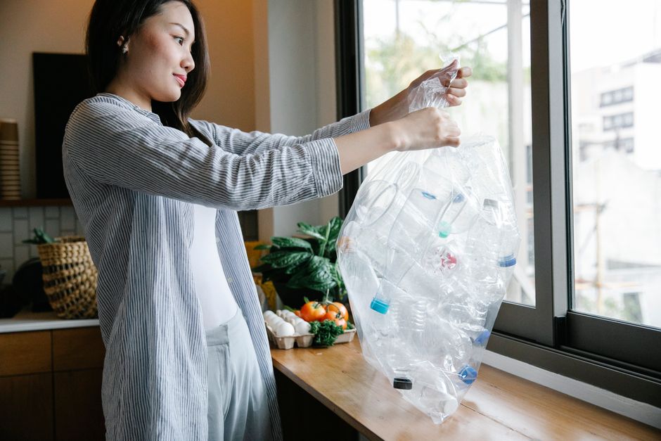A woman with long dark hair, wearing a light grey and white striped shirt and white trousers, stands inside a modern kitchen near a large window. She is holding a clear plastic bag filled with used plastic bottles and containers, preparing for disposal or recycling. Behind her on a wooden countertop, there is a black basket, a bunch of fresh vegetables including tomatoes and leafy greens, and a potted plant with large green leaves. The window reveals a blurred view of neighboring buildings and tree branches, with natural daylight illuminating the scene. The setting suggests an environment where waste sorting or recycling activities are taking place, highlighting the importance of responsible rubbish handling, possibly as part of a private collection service like those offered by Rubbish Removal Holland Park, emphasizing eco-friendly waste disposal practices inside a contemporary home.