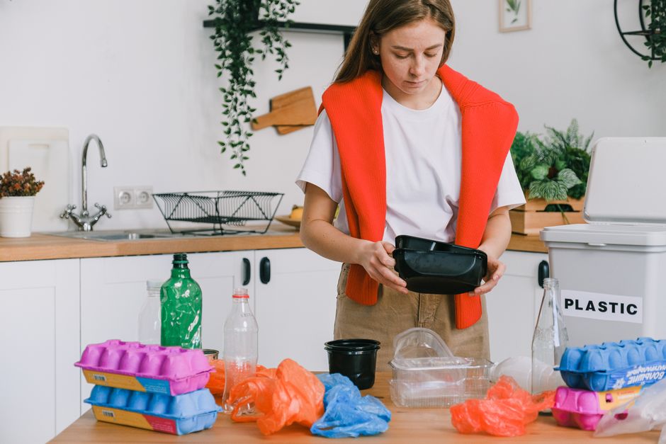 A woman with long dark hair, wearing a light grey and white striped shirt and white trousers, stands inside a modern kitchen near a large window. She is holding a clear plastic bag filled with used plastic bottles and containers, preparing for disposal or recycling. Behind her on a wooden countertop, there is a black basket, a bunch of fresh vegetables including tomatoes and leafy greens, and a potted plant with large green leaves. The window reveals a blurred view of neighboring buildings and tree branches, with natural daylight illuminating the scene. The setting suggests an environment where waste sorting or recycling activities are taking place, highlighting the importance of responsible rubbish handling, possibly as part of a private collection service like those offered by Rubbish Removal Holland Park, emphasizing eco-friendly waste disposal practices inside a contemporary home.