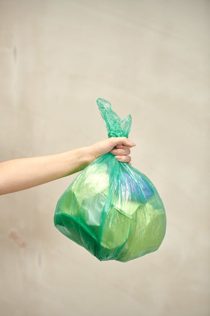 A human hand extends into the frame, gripping a green plastic rubbish bag tightly by its twisted, knotted tie, which is also made of plastic. The bag appears semi-transparent, revealing indistinct contents inside, and has a smooth, glossy surface that reflects light. The background is plain and neutral, with a soft beige or light grey tone, providing a neutral environment that emphasizes the bag and hand. The composition suggests an act of rubbish disposal or waste collection, relevant to private waste handling services such as those offered by Rubbish Removal Holland Park. The scene captures a moment of waste being prepared for removal, aligning with themes of rubbish removal and responsible waste management, subtly supporting on-site clearance or alternative disposal methods outside of traditional council services. The lighting is diffuse, with no strong shadows, creating a clear and detailed view of both the hand and the plastic bag, suitable for accessible imagery describing waste collection actions.