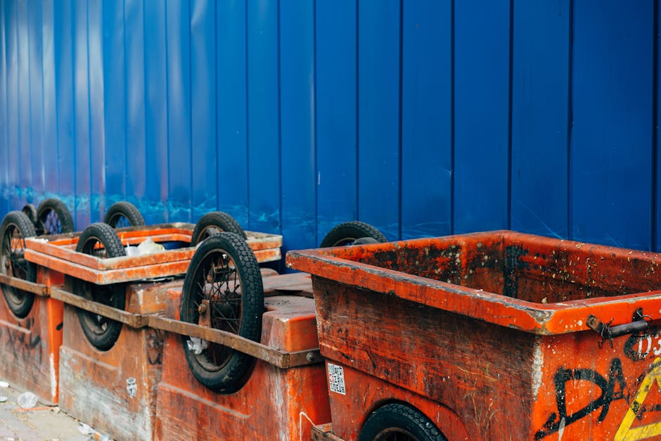 A row of orange wheeled skips positioned along a surface next to a tall, solid blue metal fence. The skips are made of heavy-duty plastic with a textured finish, showing signs of dirt, grime, and paint splatters, indicating frequent use for waste collection. The front skip has visible graffiti markings and rust patches, with a black metal latch on the side. Several black rubber tires with textured treads are mounted on the top edges of the skips for ease of movement, with some tires slightly tilted or leaning. The area appears to be an outdoor waste storage or collection point, with minimal natural light suggesting an overcast day, and the scene is clean but utilitarian, reflecting typical features of private rubbish handling infrastructure often seen in waste management services such as those provided by Rubbish Removal Holland Park. The environment suggests an emphasis on on-site waste containment, possibly as an alternative to traditional council-based rubbish collection, supporting the importance of independent waste disposal solutions.