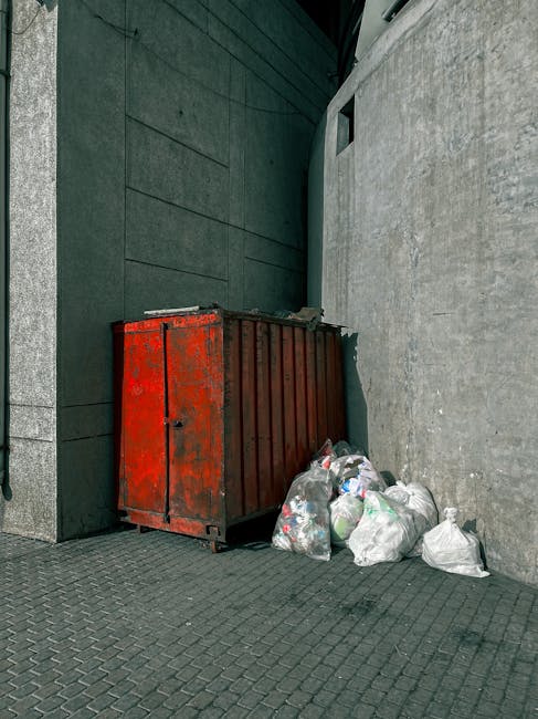 A large, red metal waste skip with a weathered and slightly rusted surface, positioned against a dark tiled wall on the left and a rough light grey concrete wall on the right. The skip is situated on a paved street with small, interlocking bricks visible in the foreground. Several white and transparent plastic rubbish bags, some loosely filled and bulging with household waste, are placed on the ground next to the skip, with a few bags leaning against it. The environment appears to be an urban alleyway or side street, with the restricted space emphasizing the collection of waste for private disposal or alternative rubbish removal services. The lighting is natural and diffused, indicating an outdoor setting without direct sunlight, and the overall scene highlights typical waste management practices involving on-site clearance in a city environment, possibly managed by a service like Rubbish Removal Holland Park.