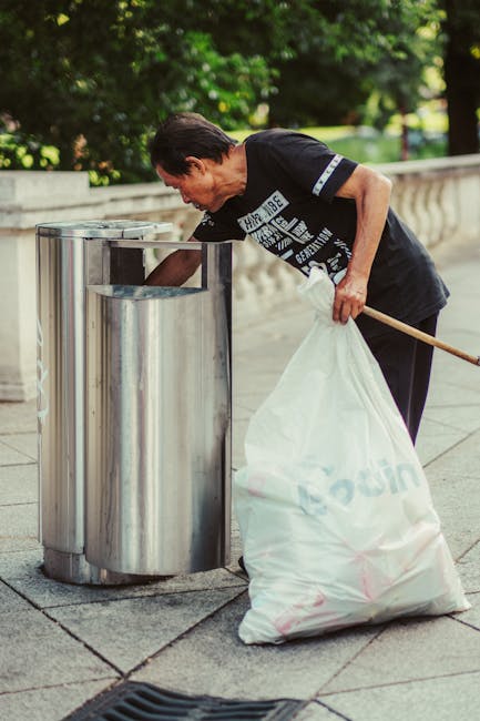 A middle-aged woman with short dark hair, dressed in a black T-shirt with white and blue text, is seen disposing of waste into a metal recycling bin located on a paved surface outdoors. She is bending forward, holding a large white plastic bag filled with rubbish in her left hand, while her right hand extends toward the open-top bin. The bin appears to be part of a row of similar recycling containers, situated in a public park or urban outdoor space with lush green trees and foliage in the background. The scene is illuminated by natural daylight, highlighting the reflective surface of the metal bin and the textured stone pavement beneath her feet. The woman is engaged in an act of waste disposal, possibly related to private rubbish collection or local rubbish removal services, which Rubbish Removal Holland Park provides as part of their rubbish clearance after events or general waste management. The environment suggests a clean, organized setting for rubbish disposal, emphasizing the importance of proper waste handling in public spaces.
