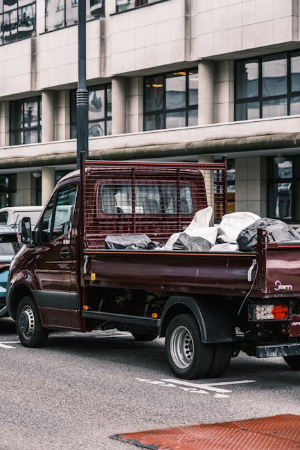 A small, dark red flatbed truck parked on an urban street in front of a modern office building with large glass windows. The truck's cargo area is filled with various types of rubbish, including black and white plastic bags and loose debris, indicating it is in the process of waste collection or disposal. The truck features a metal safety grille partition behind the cab and is situated on a paved surface with lane markings visible nearby. The scene reflects a typical private waste removal activity, likely coordinated by Rubbish Removal Holland Park, suitable for on-site clearance or alternative waste handling services in a city environment. The background shows a lamppost and additional parked vehicles, suggesting an active commercial or office district.