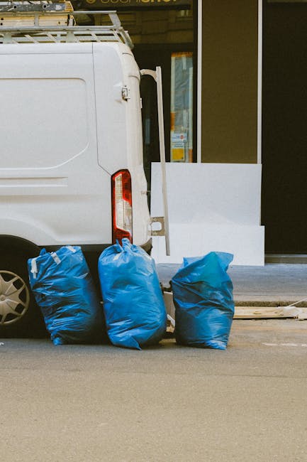 A pile of mixed rubbish consisting of black plastic garbage bags, a yellow plastic container, a worn beige car seat, and various smaller debris is gathered on a gravel surface in an outdoor area. The rubbish is positioned against a rough stone wall, with some items leaning against it, and is partially shadowed by a nearby metal pole. In the background, there is a chain-link fence topped with greenery, and beyond that, a tennis court or sports facility with a covered structure, visible through a partly cloudy sky. The scene appears to depict a typical instance of waste left for collection or illegal dumping, a situation that Rubbish Removal Holland Park may address through private on-site clearance or alternative disposal methods, reflecting the importance of professional waste management services in urban environments.