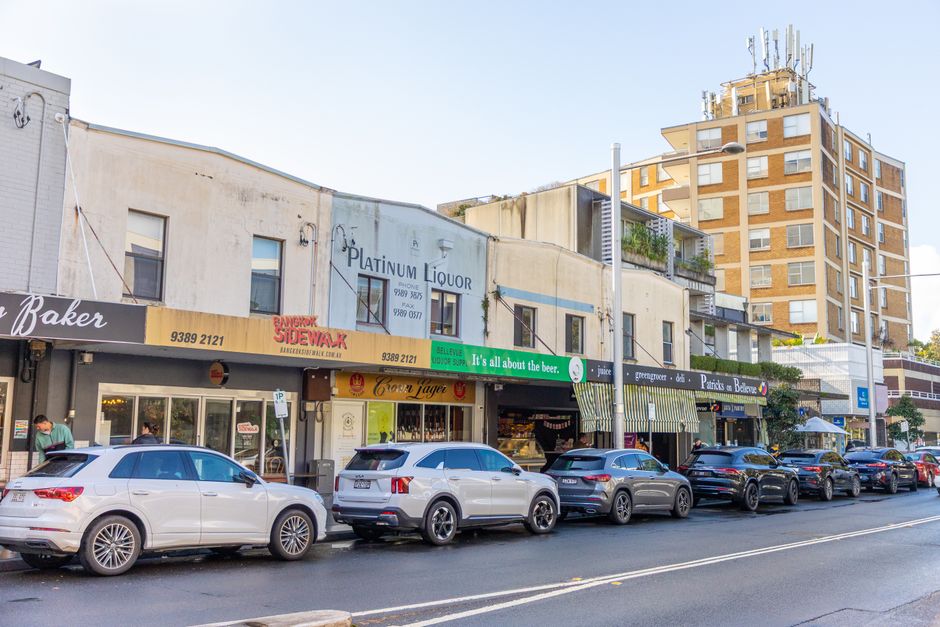 A row of commercial buildings along a street in an urban area, featuring various storefronts, including a bakery, a liquor store named Platinum Liquor, and a bar with signage indicating delivery services. The buildings have a mix of aged concrete and painted facades, some showing signs of weathering and minor wear. Several cars are parked in front, including a white SUV, a grey SUV, and multiple other vehicles stretching along the curb, emphasizing street-level parking commonly associated with business districts. In the background, a multi-story residential or office building rises, with antennas and communication equipment on its rooftop. The scene is illuminated by natural daylight, with a clear sky overhead, and a lamppost stands in the foreground, partially obstructing the view of a few storefronts. This environment, with its combination of parked vehicles and nearby shops, subtly aligns with the context of private waste handling and rubbish removal services, such as those offered by Rubbish Removal Holland Park, supporting efficient on-site clearance and alternative rubbish collection in urban commercial zones.