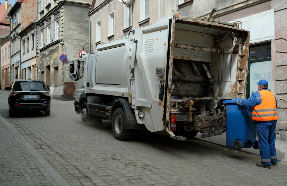 A white waste collection vehicle with an open rear loading compartment is parked along a narrow cobblestone street lined with multi-story residential buildings. The truck appears to be in the process of rubbish collection, with the compartment showing signs of rust and wear, indicating frequent use for waste handling. A worker dressed in a blue uniform and high-visibility orange vest is standing on the right side of the vehicle, actively pushing a blue wheeled rubbish bin towards the open compartment, ready for emptying. The street features parked cars, including a dark SUV on the left, and the environment suggests an urban area with older, stone-fronted buildings that have small windows and varied facades. Overhead, the lighting appears natural, typical of daytime, with soft shadows cast on the scene. The overall setting emphasizes a routine rubbish collection operation, reflecting private waste disposal services like those provided by Rubbish Removal Holland Park as an alternative to local authority rubbish collection, with a focus on on-site or private waste handling in a residential setting.
